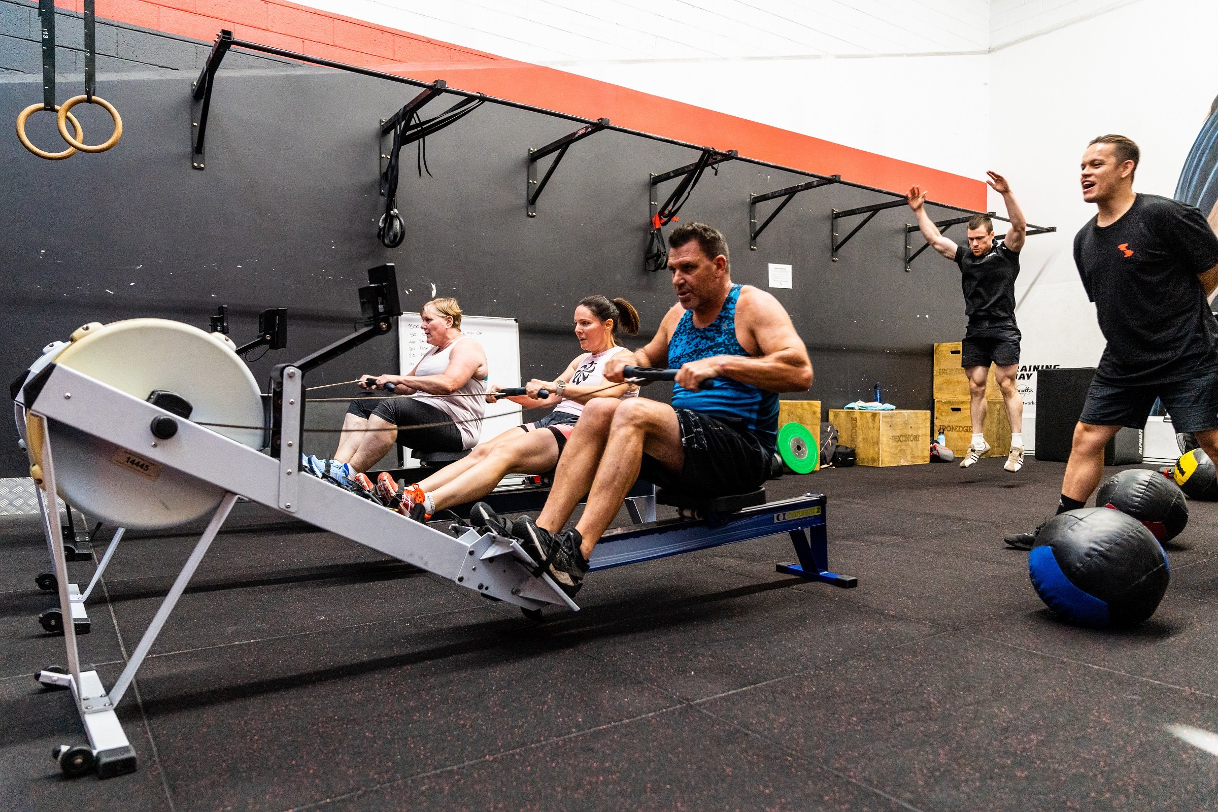People exercising in a gym doing advanced body fitness training. Two women and one man are using rowing machines, while two men in the background are engaged in other exercises.