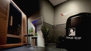 Modern recovery room with a wooden sauna, a black Primal Ice cryotherapy chamber, a round table with plants, a large mirror, and a palm plant. "Training Day Recovery" sign on the wall.