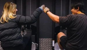 Two people sit facing each other on gym equipment, reaching out for a fist bump. One wears a black jacket and the other a dark t-shirt and shorts. The setting appears to be a fitness center or gym.